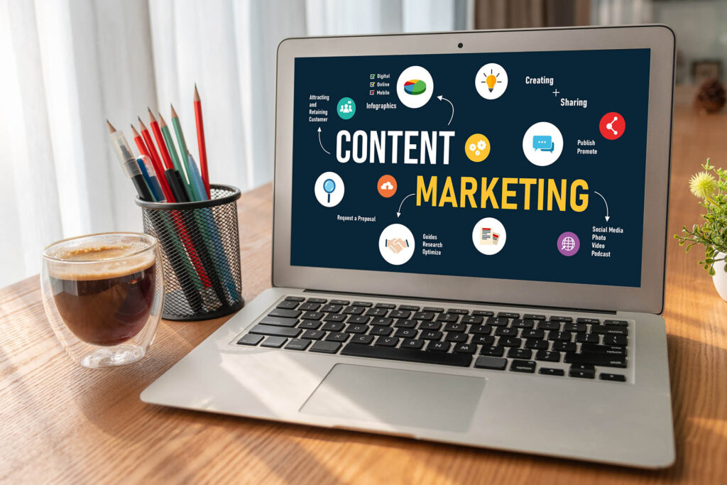 Laptop with the words “content marketing” on the screen next to a coffee cup, pens and pencils, and a potted plant on a wooden surface.