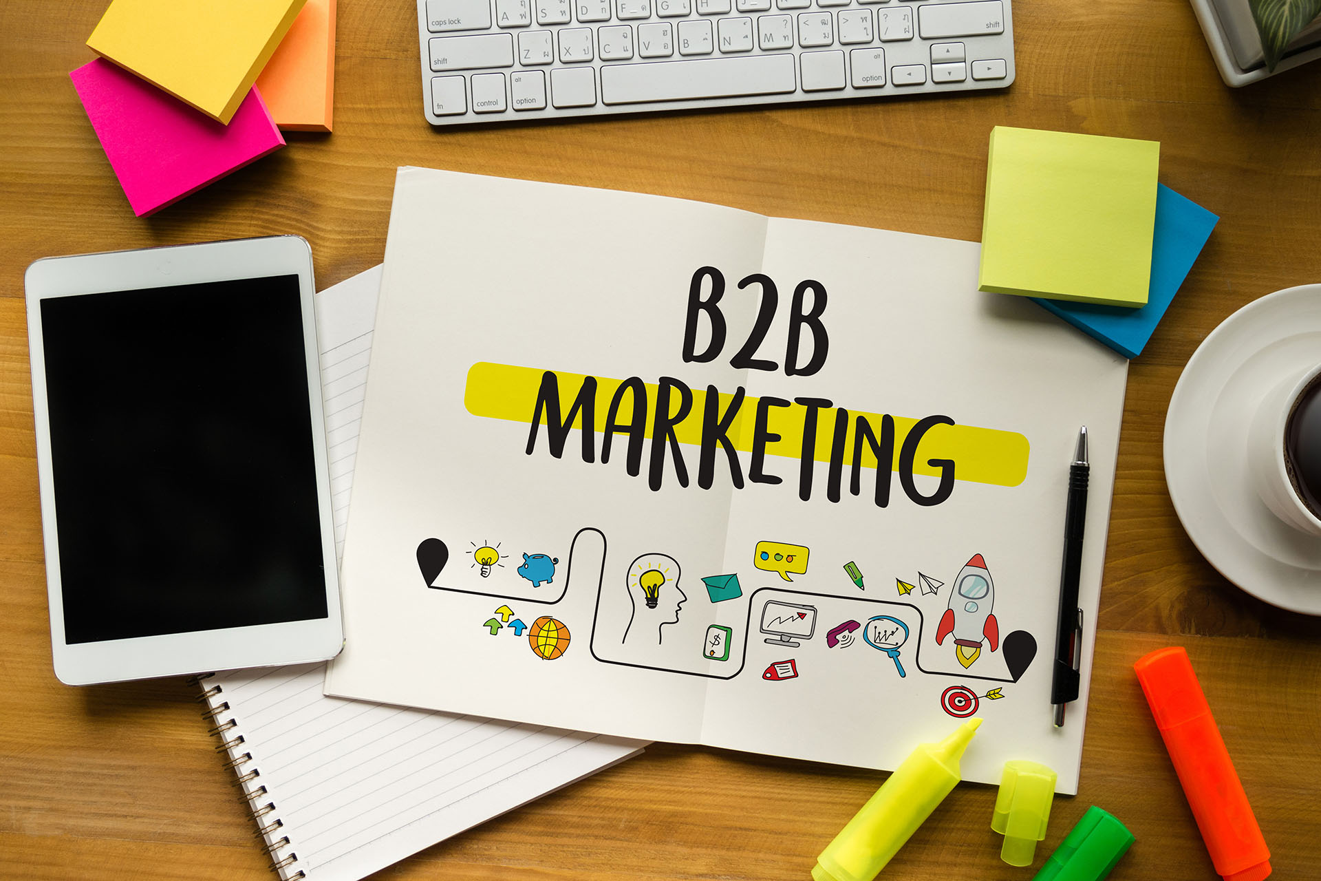 “B2B Marketing” on a paper with icons, surrounded by pens, highlighters, a notebook, and iPad, a coffee cup, sticky notes, and a keyboard.