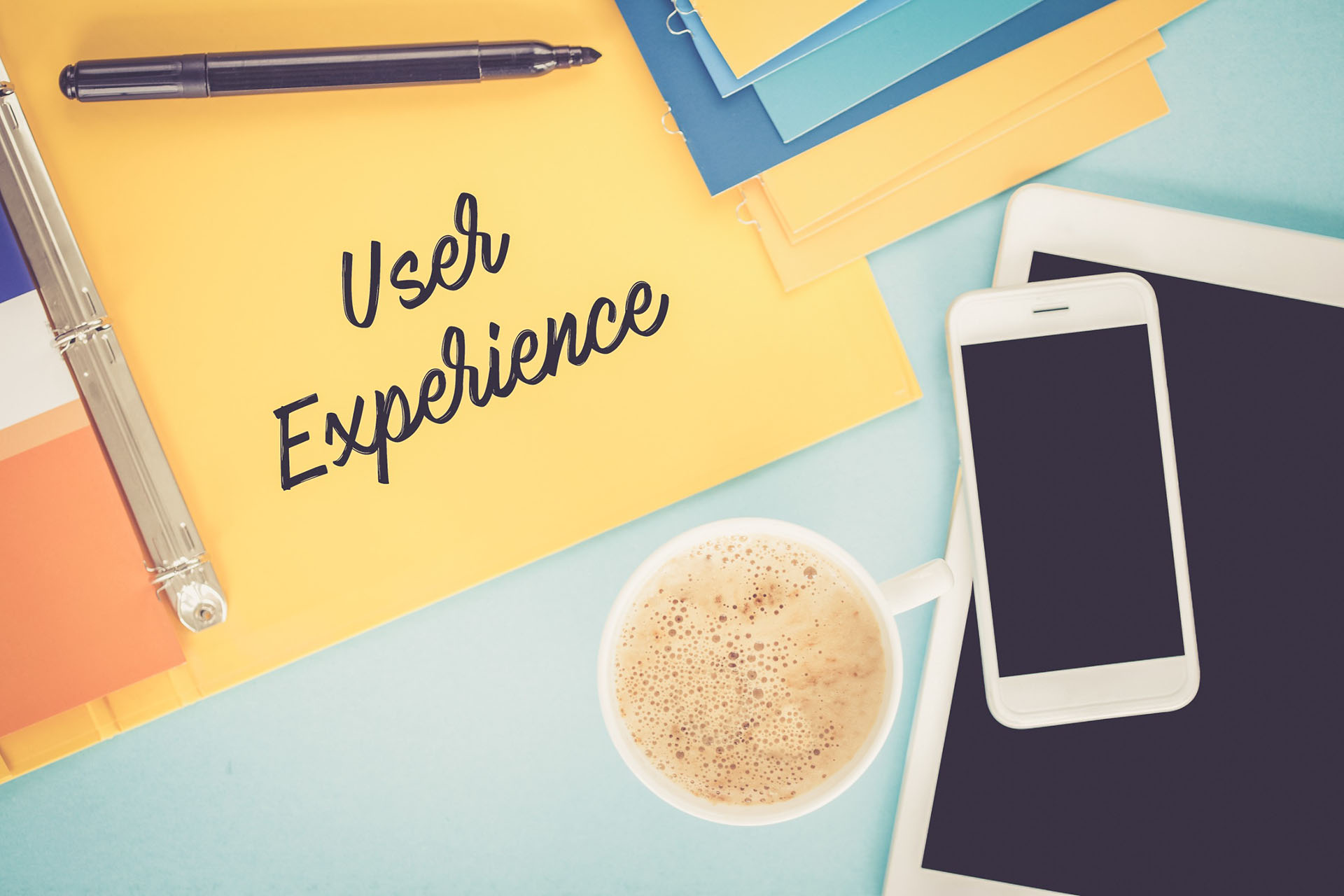 Words “user experience” written in cursive on a binder next to a cup of coffee, a phone, and an iPad.