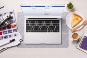 Laptop with the Facebook log in screen next to a camera, photos, a piece of pizza and a fork on a plate, a plant, a cup of coffee, and a notebook