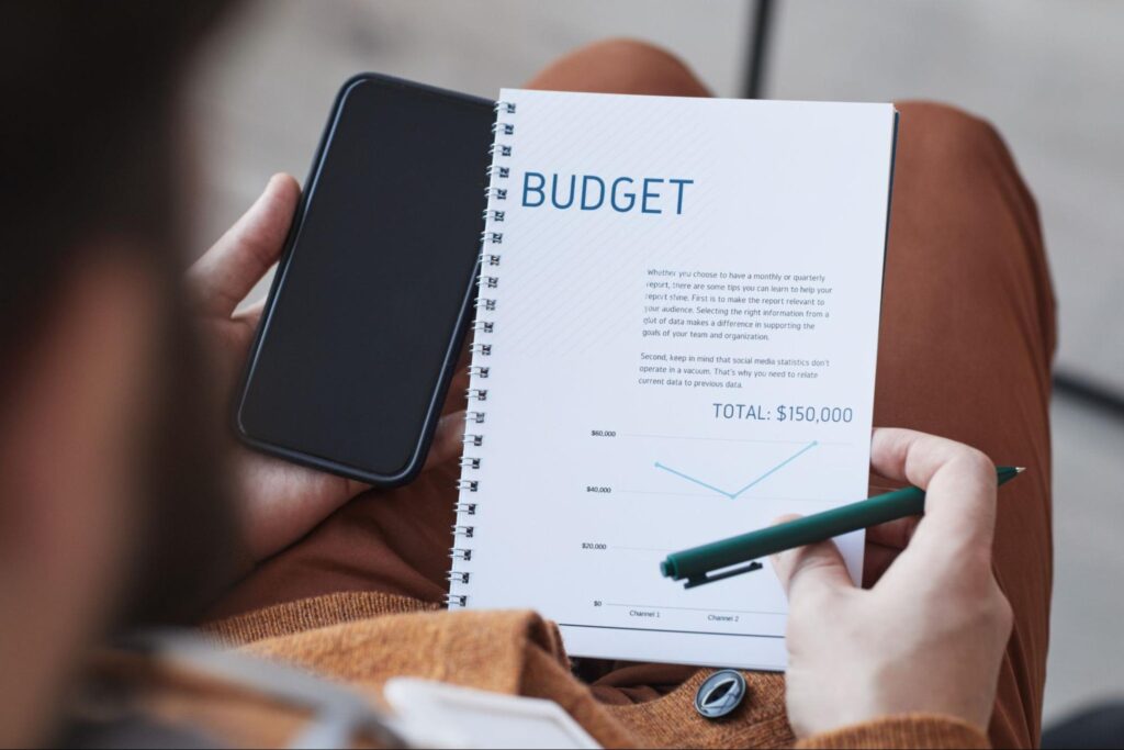 Person holding a phone in one hand and a pen in the other with a notebook in their lap that has the word budget in a large font.