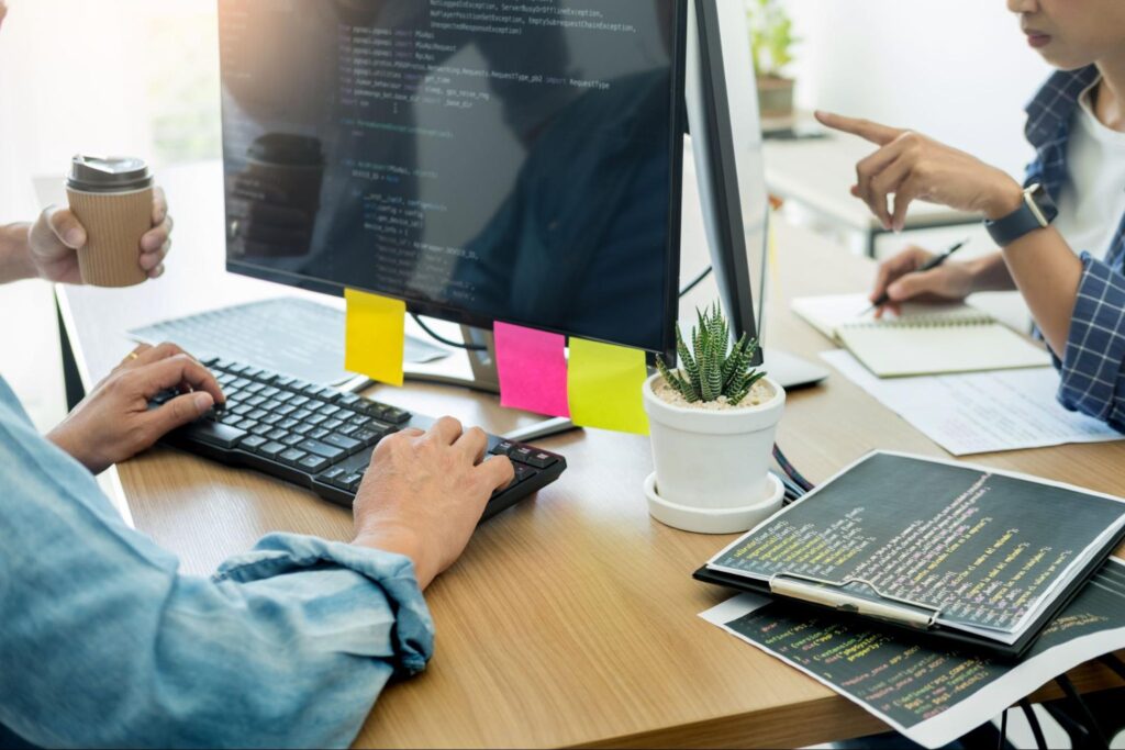 Person typing on a computer on a wooden desk with sticky notes on the screen and a potted plant beside with two people in the background.